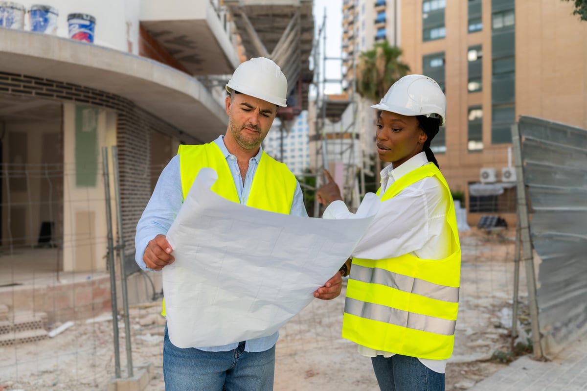 Two engineers wearing safety vests and helmets reviewing building plans beside a modular construction site